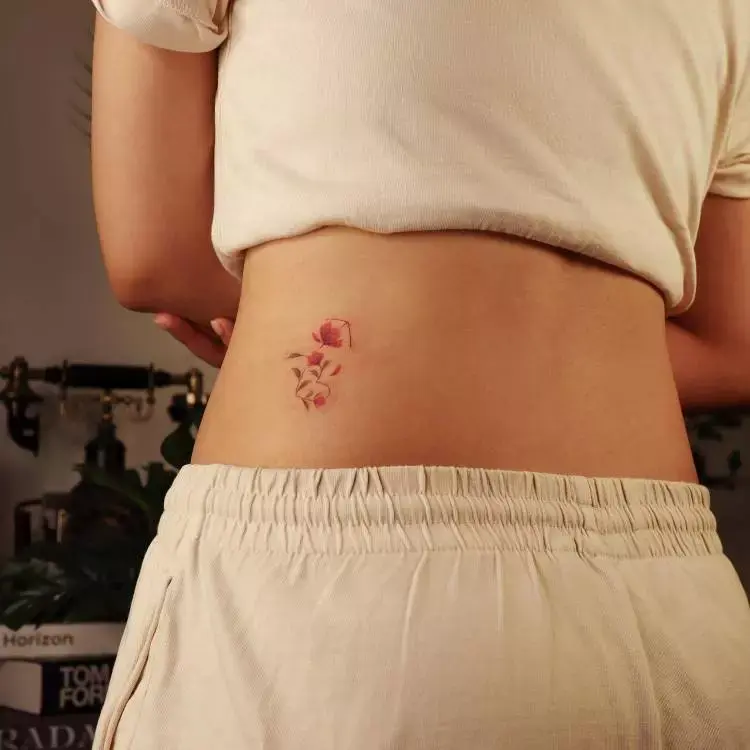 A imagem mostra uma pessoa de costas, vestindo uma camiseta e calça bege. No lado direito das costas, há uma pequena tatuagem delicada de flores em tons de rosa e verde. A cena tem um fundo desfocado com plantas e livros, criando um ambiente aconchegante e tranquilo.


