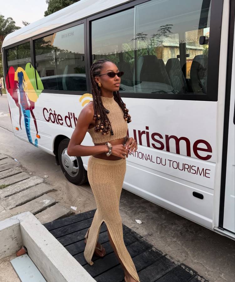 Uma mulher elegantemente vestida em um conjunto de tricô bege, saia com fenda e top cropped, caminha ao lado de um micro-ônibus branco com a inscrição "Côte d'Ivoire Tourisme", sob a luz do sol.