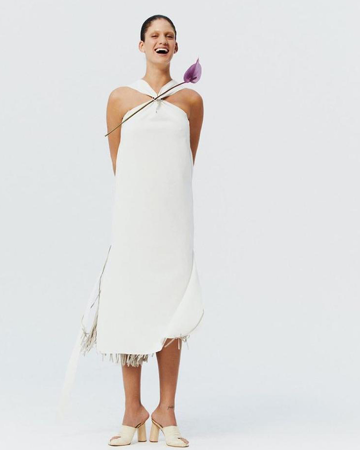 Modelo usa vestido branco sem mangas com flor roxa.
