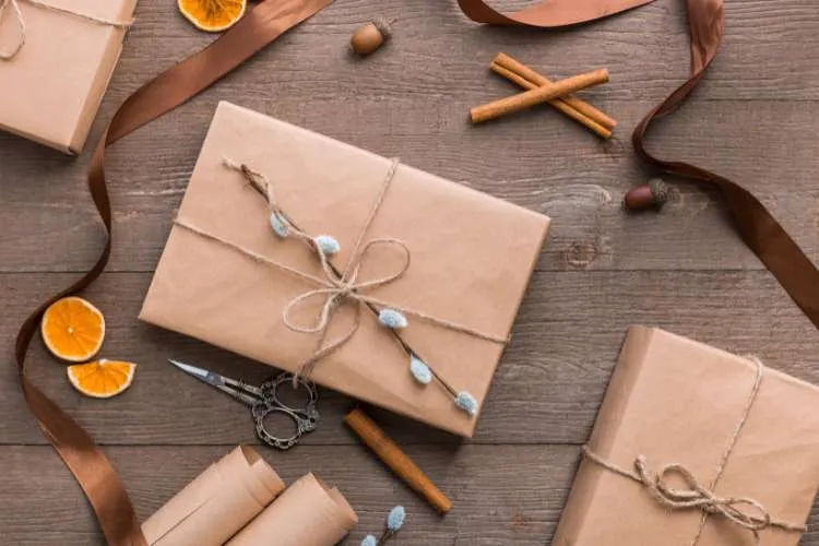 Embrulho de presente com papel kraft em uma mesa com outros embrulhos, canela, laranja e tesoura.