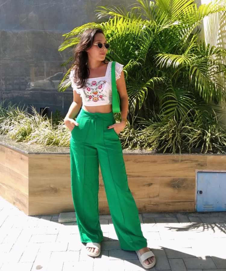 Imagem com fundo de área externa, jardim com plantas. No centro, mulher usando cropped branco + calça verde + bolsa verde