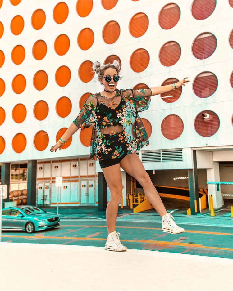 Mulher loira de pele clara com cropped e shorts pretos, com blusa transparente de flores bordadas
