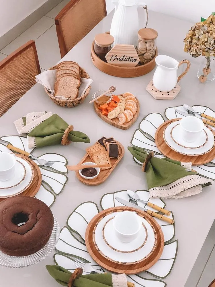 Mesa posta em tons de marrom e verde com bolos e pães. O suplat é branco em formato de Monstera