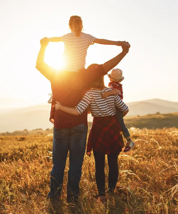 Família de costas em um campo dourado ao pôr do sol. O pai leva a filha mais velha nos ombros e a mãe, a mais nova. A luz do sol brilha entre eles, simbolizando união e esperança.