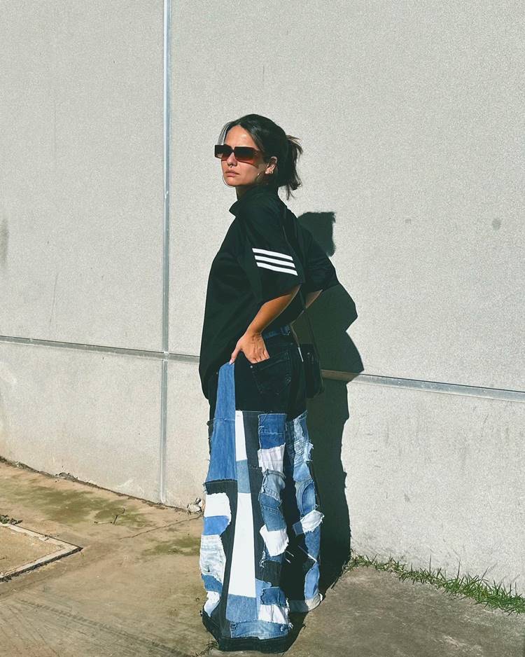 Mulher usando camiseta preta de time com calça jeans patchwork, óculos de sol e brincos de argola, em local com parede de cimento.