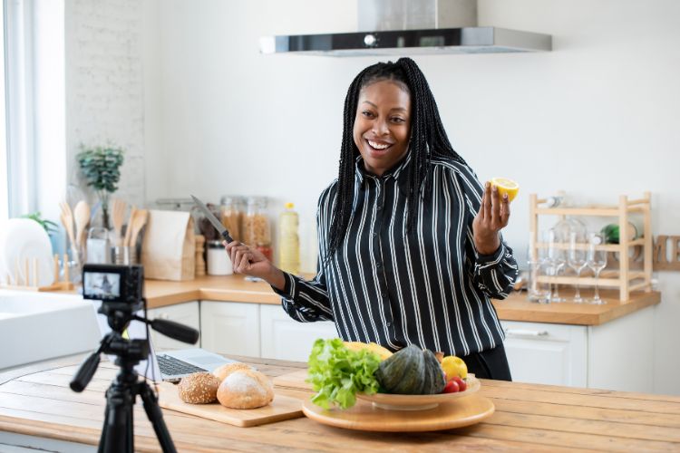 Uma mulher sorridente, com cabelo trançado, grava um vídeo de culinária para a **faculdade de influencers**. Ela segura uma faca e um limão, diante de uma câmera em tripé, com vegetais e pães sobre a mesa da cozinha.