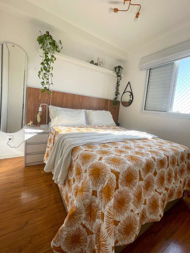 Quarto com cama de casal com jogo laranja e prateleira alta com vaso de jiboia pendente.