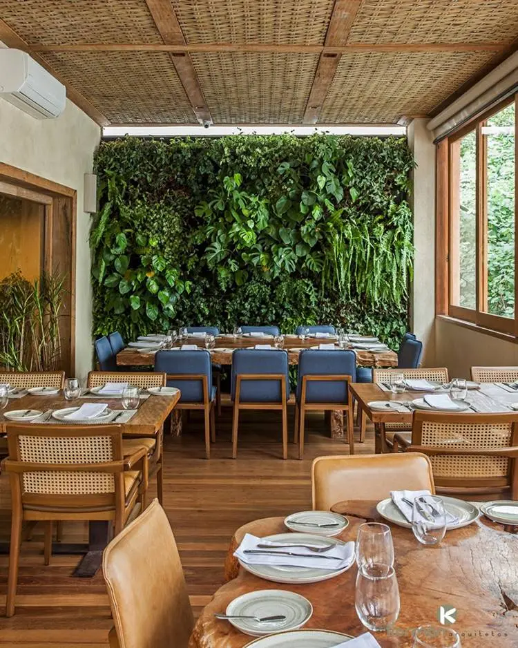 Jardim vertical verde em frente a mesa com cadeiras azuis e outras mesas provavelmente de um restaurante ou salão de festas