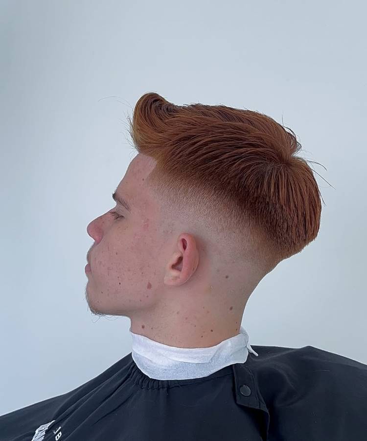 Um jovem de perfil com um corte de cabelo mid fade e pompadour. O cabelo é ruivo/acobreado e volumoso no topo, penteado para a frente e para cima. As laterais e a nuca têm um degradê.







