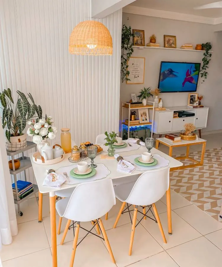 A imagem mostra uma sala de jantar bem iluminada e decorada de forma aconchegante. Há uma mesa retangular branca com pernas de madeira, cercada por quatro cadeiras brancas com pés de madeira. Sobre a mesa, há louças brancas com detalhes verdes, copos de vidro e guardanapos dobrados. No centro, um vaso com uma planta verde adiciona um toque de frescor.

Ao lado da mesa, há um carrinho com rodas, que contém uma jarra de suco de laranja, xícaras e um arranjo de flores brancas. Uma luminária pendente de palha ilumina a mesa.

No fundo, há uma estante branca com prateleiras, decorada com plantas, quadros e objetos decorativos. Uma televisão está ligada, exibindo uma imagem de um beija-flor. O piso tem um padrão geométrico em tons neutros, e há uma sensação geral de harmonia e conforto no ambiente.