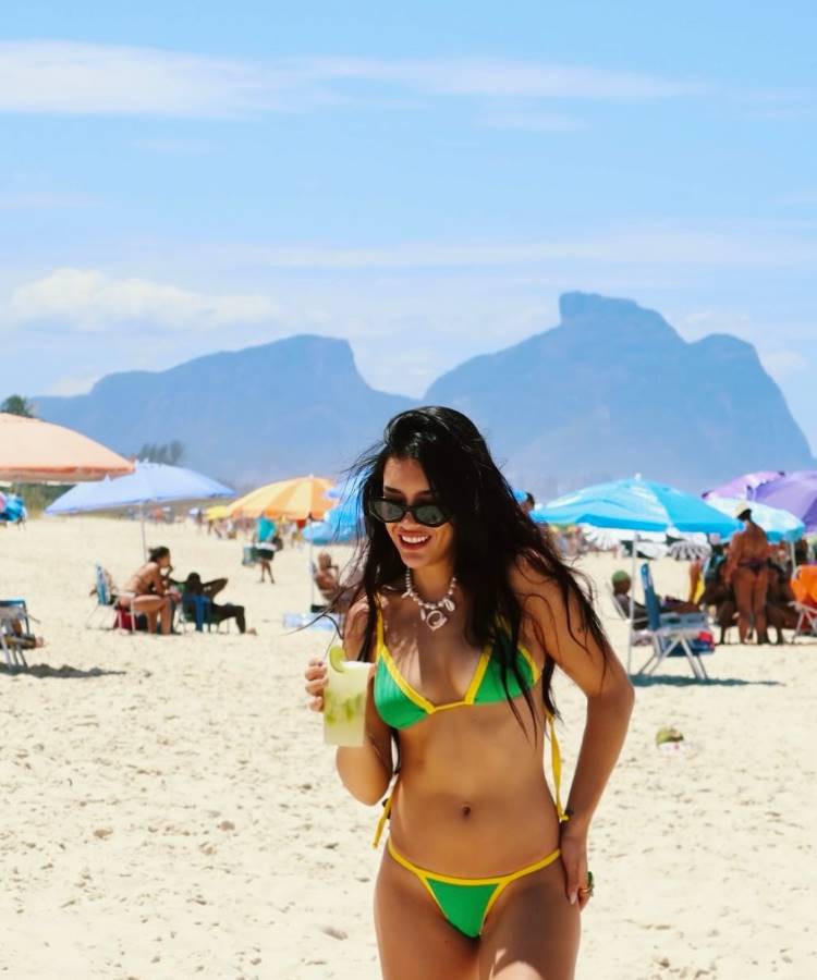 Uma mulher de biquíni verde e amarelo caminha em uma praia ensolarada, segurando uma bebida. Ao fundo, várias pessoas estão sob guarda-sóis coloridos, e há montanhas à distância, sob um céu azul claro. Ela usa óculos escuros.