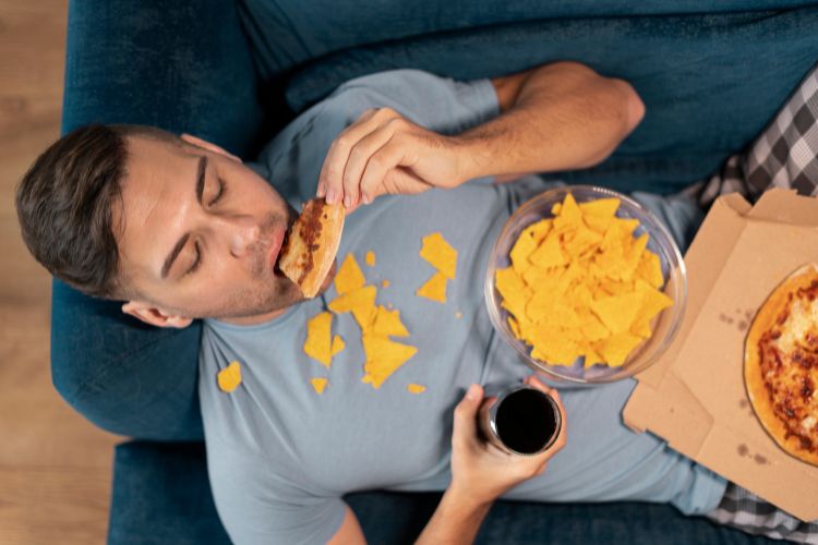 Homem deitado no sofá comendo salgadinho e pizza