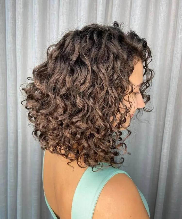 Uma mulher de perfil com um corte de cabelo bob cacheado volumoso, em tons de castanho. Os cachos são bem definidos e saudáveis. Ela veste uma blusa de alças azul-claro contra um fundo de cortinas claras.