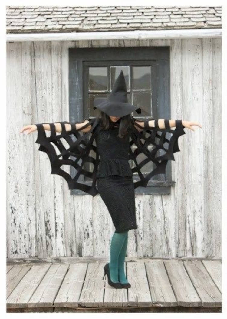 Mulher de pele clara usando chapéu de bruxa, blusa e saia pretas, poncho de teia de aranha e meia-calça verde