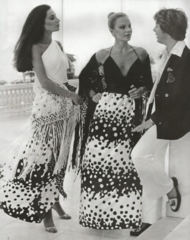 Imagem em preto e branco do estilista brasileiro Guilherme Guimarães nos anos 70 em pé a conversar com duas modelos com vestidos longos Imagem em preto e branco do estilista brasileiro Guilherme Guimarães jovem em pé a conversar com duas modelos com vestidos longos