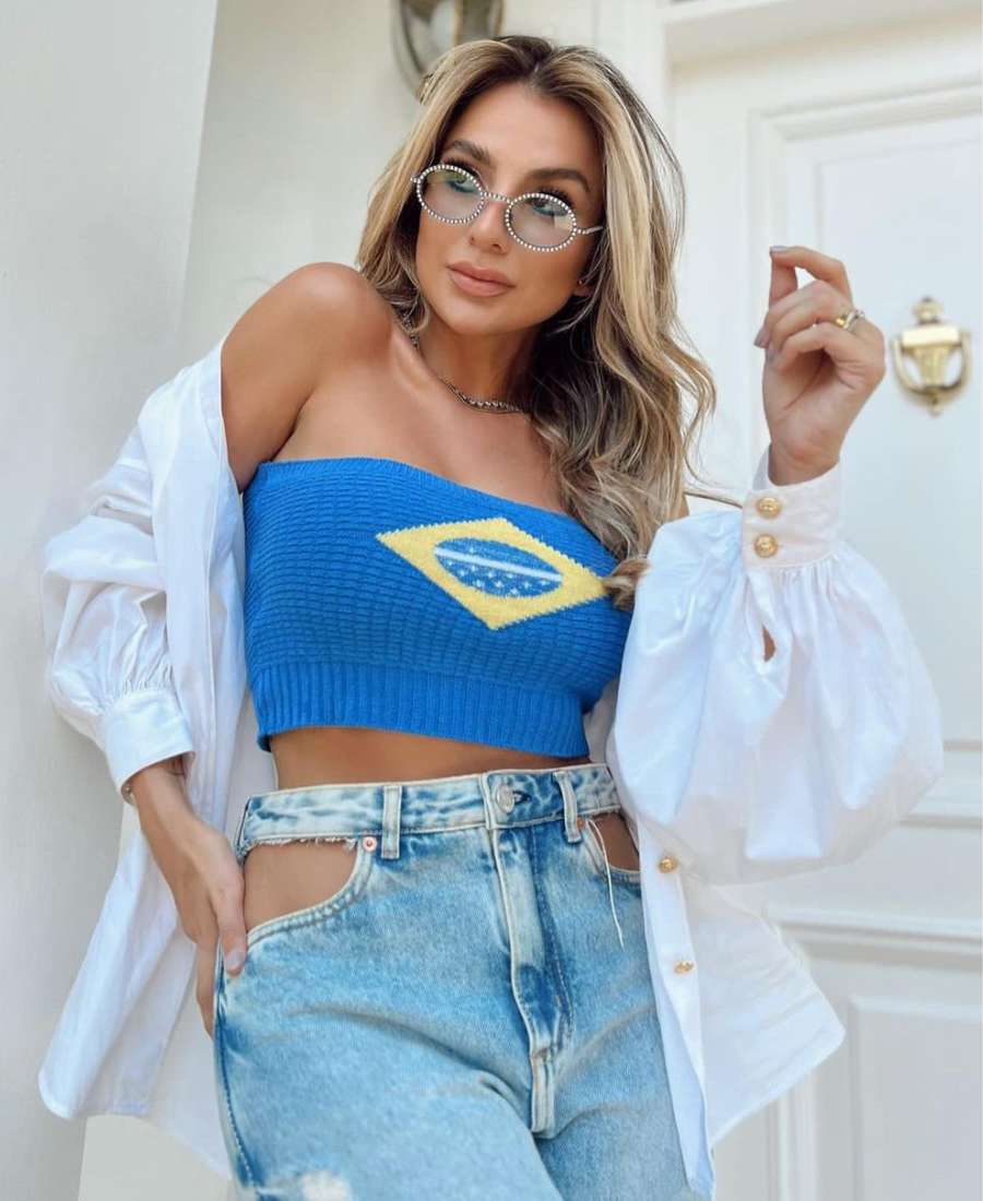 Imagem com fundo de cômodo de casa, com porta e parede. No centro, mulher usando roupas para a copa do mundo: top azul com bandeira do Brasil, calça jeans e camisa branca por cima.