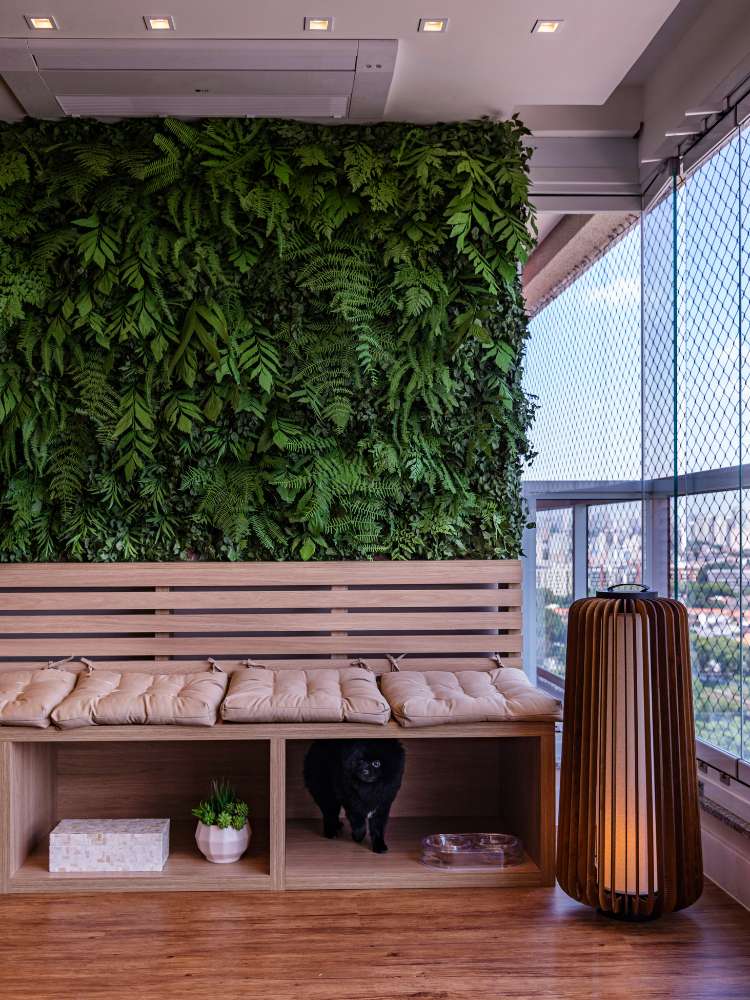 Varanda fechada com vidro, com jardim vertical e banco de madeira forrado com almofadas, além de vasos pequenos embaixo, espaço para vasilha de cachorro e luminária