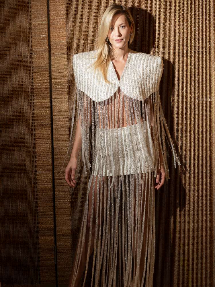 Mulher loira de pele clara usando vestido com ombreiras brancas grandes, saia branca e franjas brilhantes