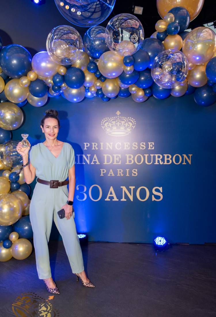 Uma mulher em macacão azul claro com cinto marrom, segura um frasco de perfume. Ela está em um evento com tema em azul e dourado, decorado com balões. A parede atrás dela exibe "Princesse Marina de Bourbon Paris – 30 anos".