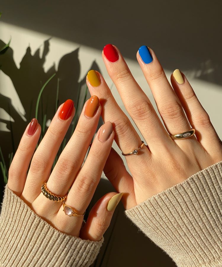 A imagem mostra duas mãos com unhas pintadas em cores vibrantes e variadas: vermelho, azul, amarelo, laranja e nude. As mãos estão adornadas com anéis finos e delicados, alguns com pequenos detalhes, como pedras. As mangas de um suéter bege cobrem parcialmente os pulsos. A iluminação natural cria sombras suaves e destaca as cores das unhas e o brilho dos anéis. Ao fundo, há uma sombra de folhas, sugerindo a presença de plantas próximas.