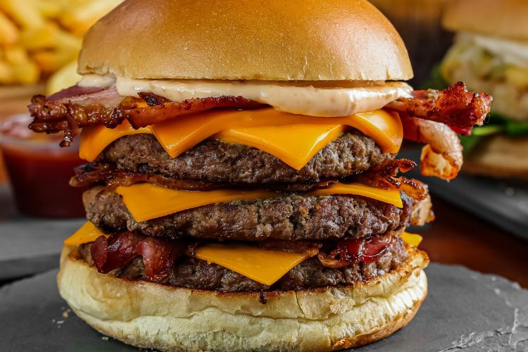 Hambúrguer suculento triplo com camadas de queijo derretido, fatias de bacon crocante e molho, servido em pão macio dourado. Ao fundo, batatas fritas e outro hambúrguer desfocados, compondo um cenário apetitoso e irresistível.