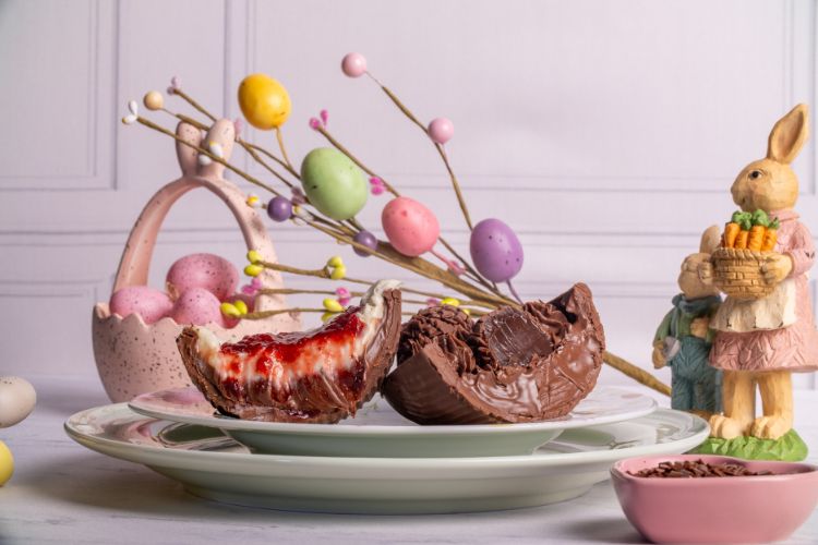 Metades de ovos de chocolate recheados com creme e geleia estão sobre um prato verde. Ao fundo, decoração de Páscoa com coelho de cerâmica, ovos coloridos em galhos e cesta com ovos rosa completam o cenário festivo.