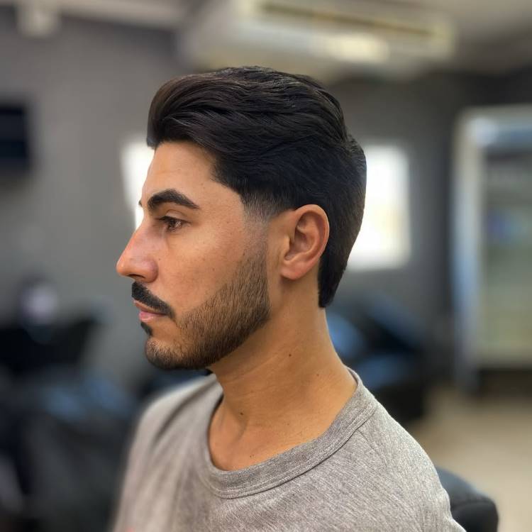 Um homem com bigode e barba, de perfil. Ele tem um corte de cabelo pompadour com volume no topo, e as laterais e a nuca têm um mid fade com degradê sutil.