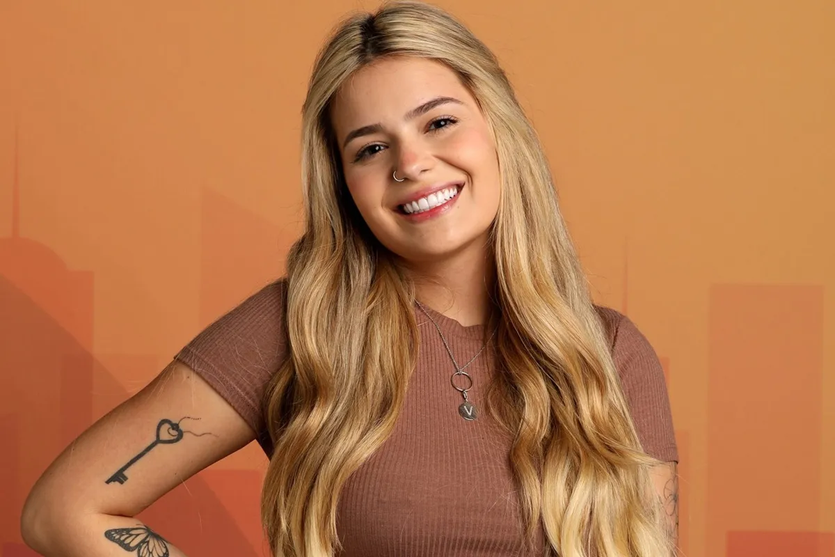 Retrato de uma participante do Big Brother Brasil sorrindo, com fundo em tom alaranjado, em imagem promocional do reality show