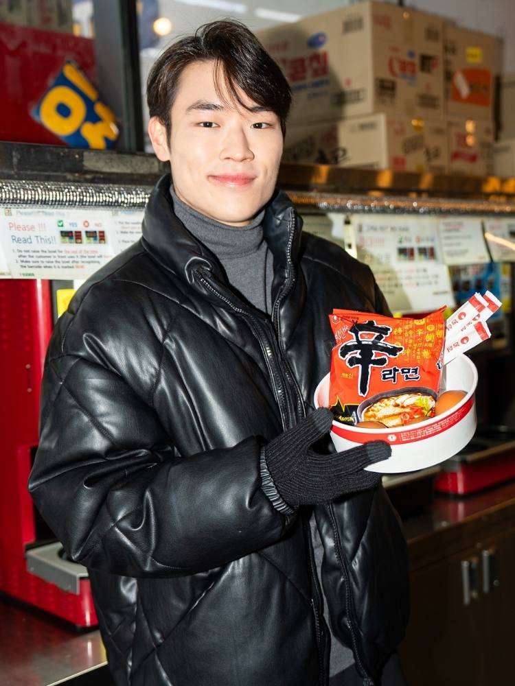 Arthur Paek usando blusa cinza de gola alta, jaqueta preta de couro e segurando embalagem de macarrão coreano