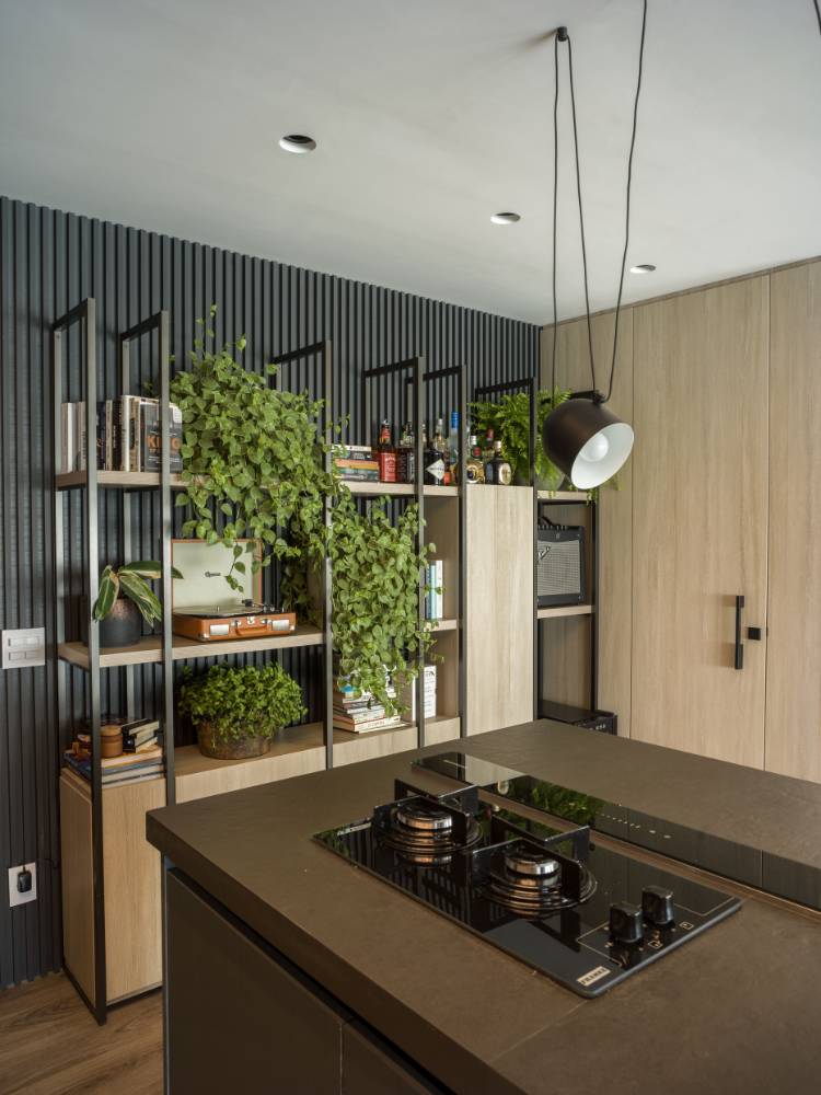Estante com nichos abertos e fechados, colocada a frente de um painel ripado preto. Ela pertence ao ambiente da cozinha e da sala. Nela, há plantas, plantas, quadros, bebidas, livros e até vitrola. 