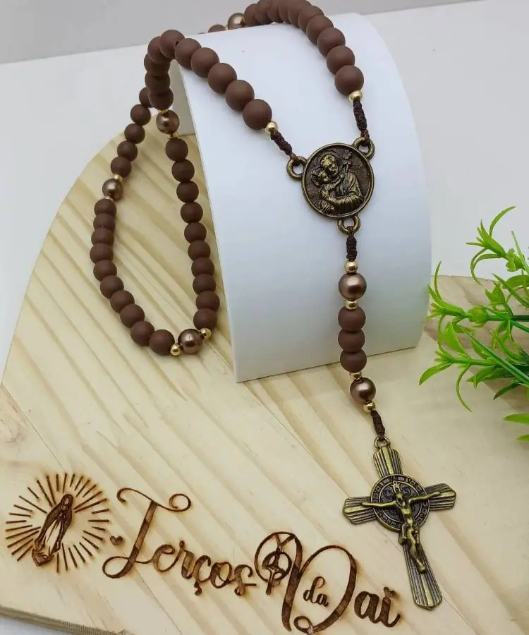 A segunda imagem mostra um terço marrom com contas foscas e metálicas, um medalhão com uma imagem de santo e um crucifixo trabalhado. Ele está sobre uma base de madeira com a frase recortada "Terços do Pai".

