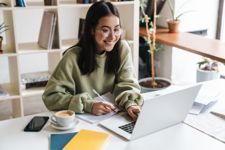 Jovem sorridente, de óculos e moletom verde, trabalha em um laptop em uma mesa com livros, caderno, celular e café. Ambiente iluminado com plantas ao fundo. Ela verifica conteúdo usando um verificador de plágio para garantir autenticidade.