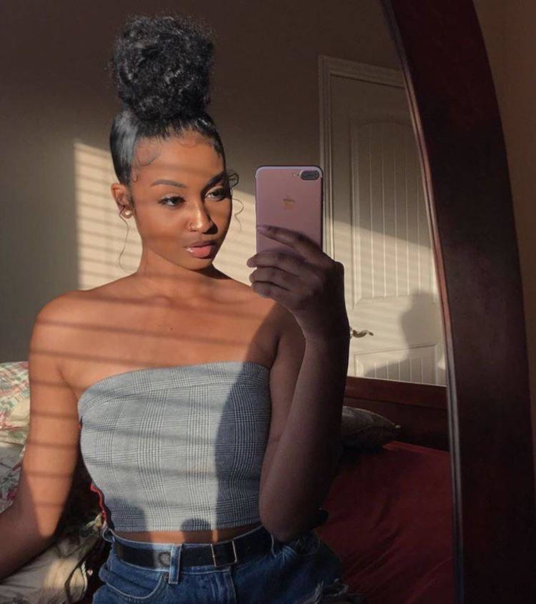 Mulher de pele negra em foto no espelho usando top cinza, shorts jeans e cabelo preto em coque volumoso