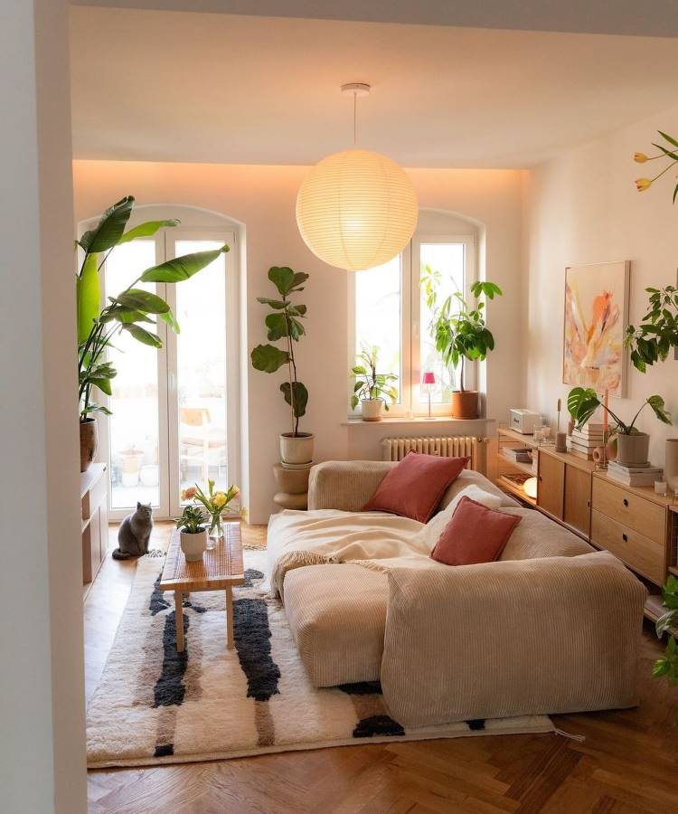 Sala de estar com decoração Feng Shui, composta por sofá bege com almofadas terracota, luz natural abundante, plantas vibrantes, mesa de madeira, tapete texturizado e luminária redonda pendente. Espaço organizado promove harmonia, energia equilibrada e um ambiente acolhedor.