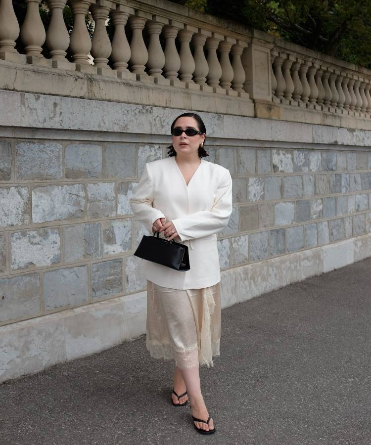 Uma mulher com óculos de sol, vestindo um blazer branco e uma saia de seda creme com renda, caminha por uma calçada. Ela segura uma bolsa preta de mão e usa chinelos, em frente a uma parede de pedra e um corrimão.