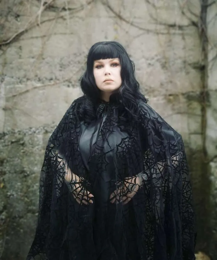 Mulher de pele clara usando vestido preto e capa de teia de aranha de tule 