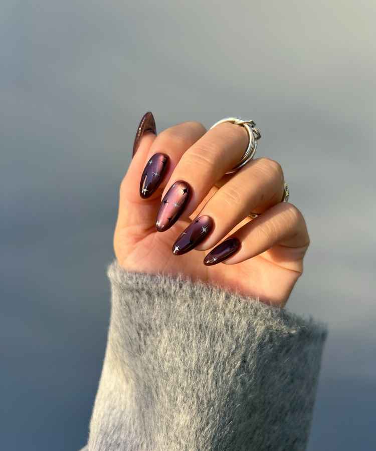 Unhas longas com esmalte roxo metálico, destacando pequenos detalhes de estrelas prateadas. A mão está parcialmente fechada, usando anéis prateados. O fundo é suave e neutro, enquanto uma manga de lã cinza adiciona textura e contraste ao visual elegante.