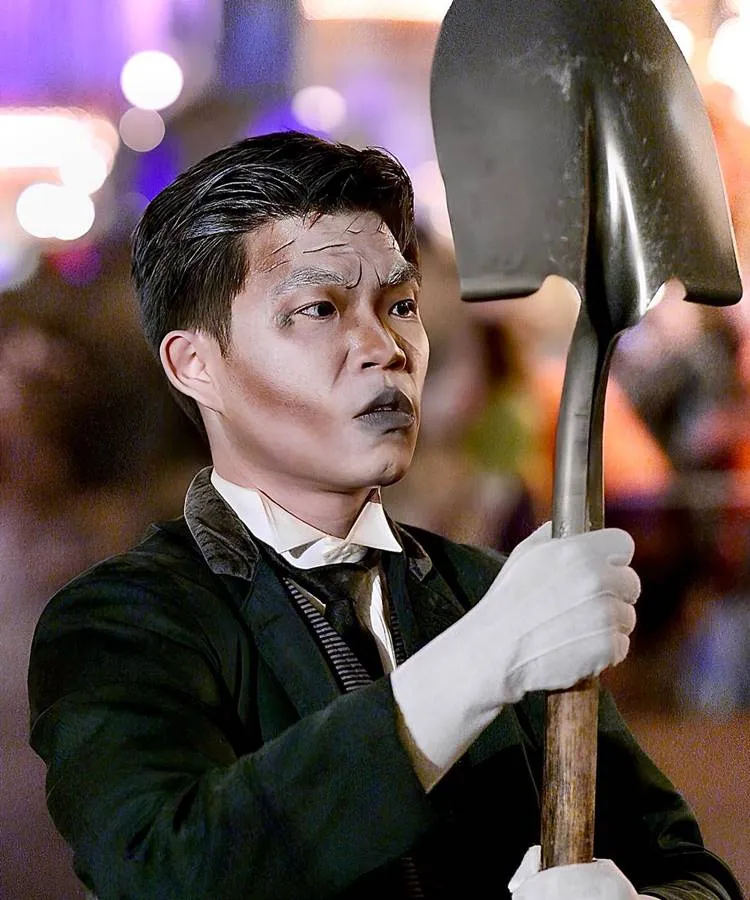 Homem asiático com maquiagem de zumbi, roupas pretas e segurando pá 