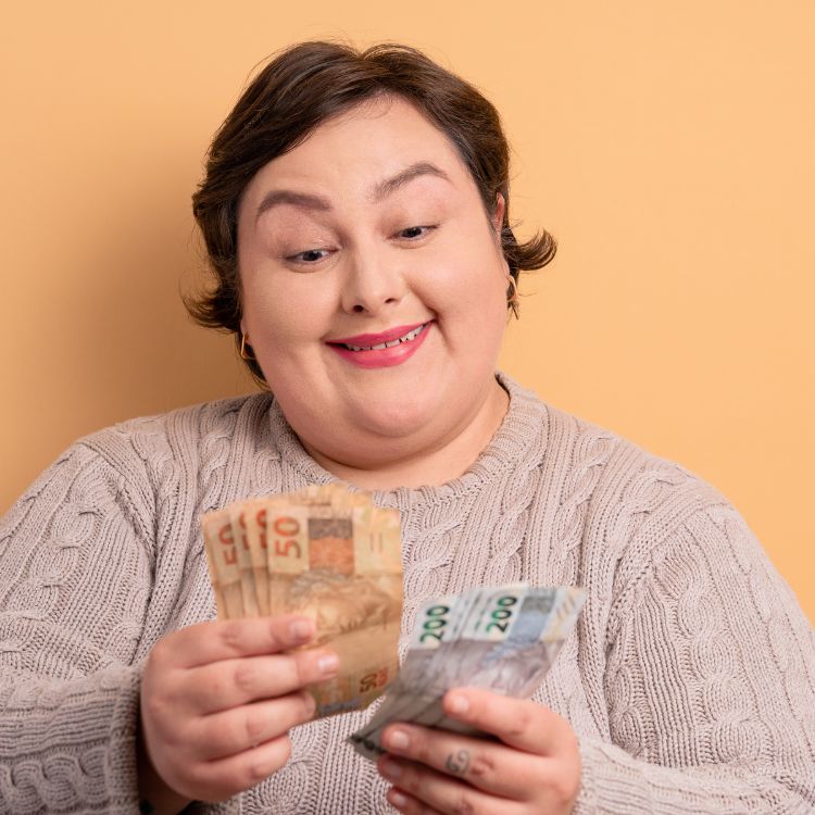 A imagem mostra uma mulher sorridente, segurando cédulas de reais (R$50 e R$200), com expressão de satisfação. Vestindo uma blusa bege, ela está em frente a um fundo laranja, simbolizando confiança. A cena reforça a importância de investimentos blindados.