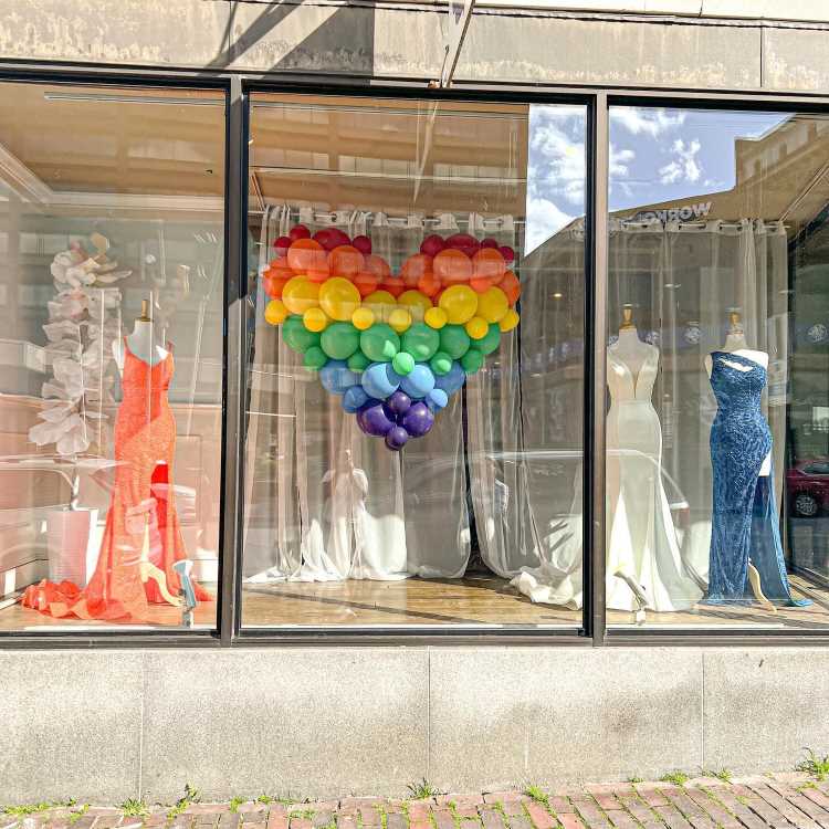 Vitrine com manequins exibindo vestidos elegantes nas cores laranja, branco e azul. Ao centro, um coração feito de balões arco-íris sobre fundo de cortinas brancas. Reflexos de edifícios completam o cenário urbano ensolarado e vibrante.