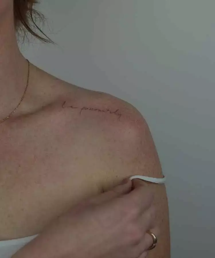 A imagem mostra uma tatuagem no ombro esquerdo, com a frase cursiva e delicada "live passionately". A pele é clara, com uma tonalidade levemente avermelhada. A pessoa veste uma alça branca simples e usa um anel dourado discreto.
