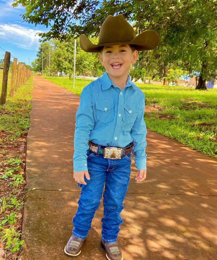 Menino usando look country infantil com camisa xadrez, calça jeans, cinto com fivela grande, sapatão e chapéu cowboy.
