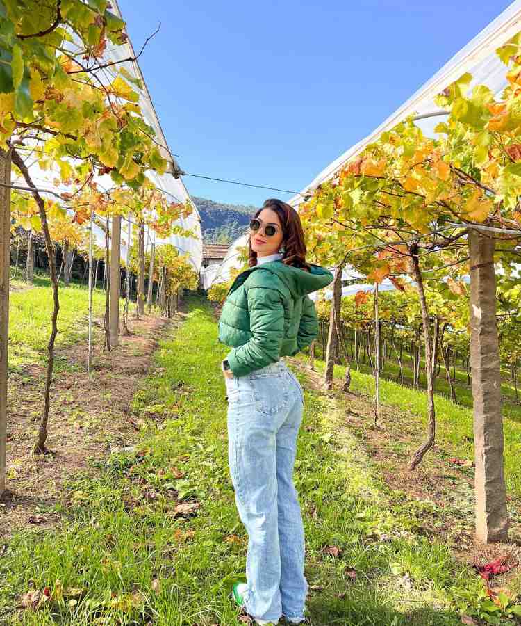 Imagem com fundo de vinhedo. No centro, mulher usando casaco puff verde + calça jeans + tênis nike verde
