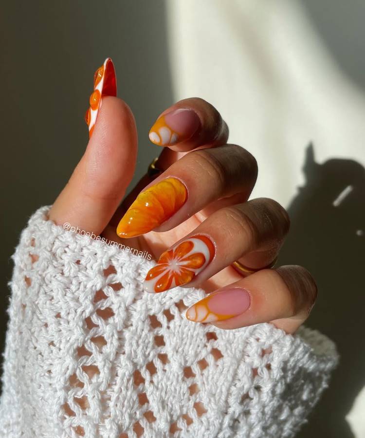 A imagem mostra uma mão com unhas decoradas artisticamente em tons vibrantes de laranja, com desenhos florais e marmorizados. A luz natural destaca os detalhes, enquanto um suéter branco de crochê contrasta suavemente com o design moderno das unhas.