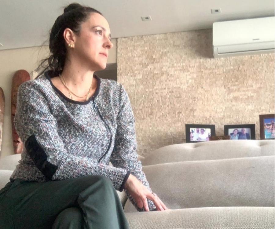 Foto de Carol Olival. Ela está sentada em sofá em uma sala. Ao fundo, parede com ar-condicionado e porta-retratos com fotografias.