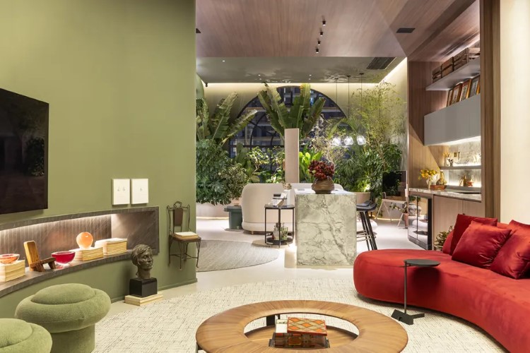 A imagem apresenta um interior moderno e elegante de uma sala de estar, com paredes verdes, sofá vermelho em forma de L, e mesa redonda de centro. Há uma TV na parede e uma área de jantar ao fundo com cadeiras estilizadas e iluminação embutida no teto. Uma grande janela mostra vegetação externa, adicionando um contraste natural ao ambiente contemporâneo.

