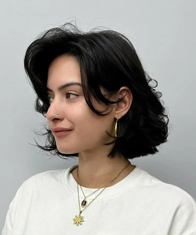 Uma mulher jovem sorri suavemente, exibindo um corte de cabelo riviera bob preto e elegante. Ela usa um brinco de argola dourado, dois colares delicados e um piercing no nariz, olhando para a esquerda da imagem.







