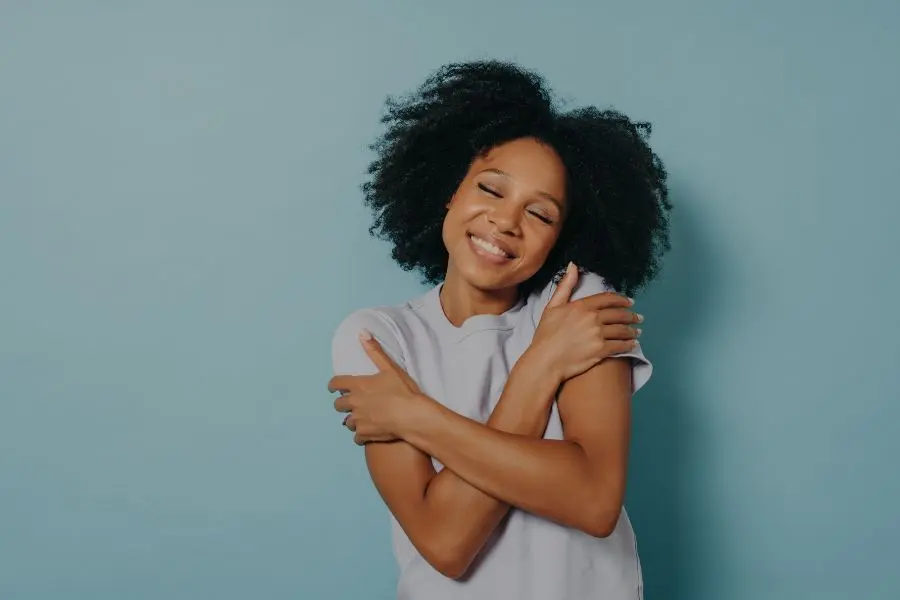 foto de mulher negra se abraçando. Ela usa camiseta branca e está na frente de uma parede azul
