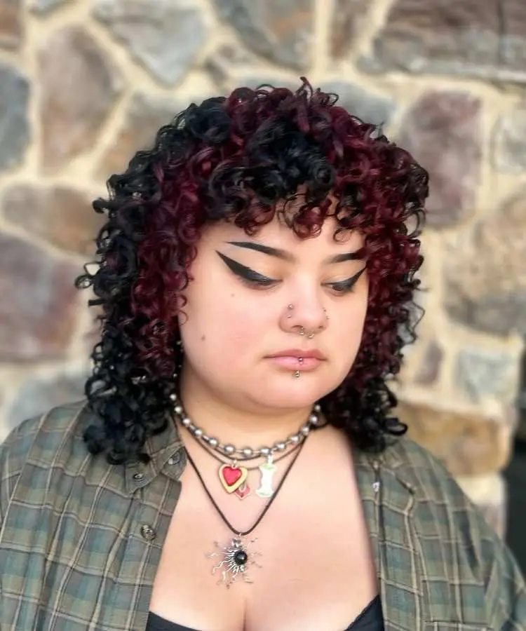 Mulher com cabelo médio cacheado, preto com mechas vermelhas, corte em camadas com franja cheia. Ela usa maquiagem marcante com delineado preto, piercings no nariz e lábio, camisa xadrez, colares variados e fundo de parede de pedras.