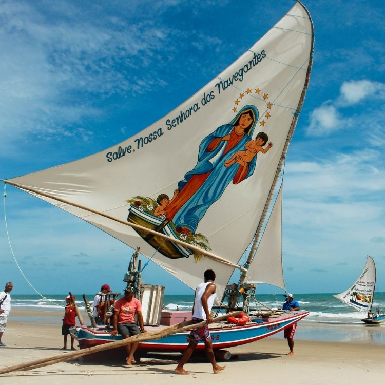 Foto de pessoas na praia com embarcação cuja vela tem desenho de Nossa Senhora dos Navegantes